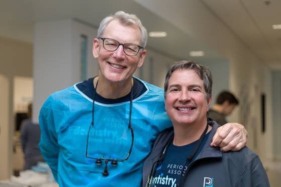 Two smiling dental professionals posing together in clinic hallway, one wearing blue scrubs and the other in dark jacket